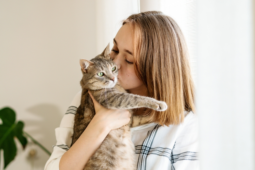 A young woman gently holds and kisses her tabby cat in a sunlit room - Pet Hospital Collierville