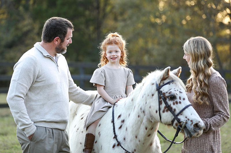 A young red-haired girl sits on a spotted horse, smiling, with a man and woman standing beside her in a grassy, tree-lined setting - Pet Hospital Collierville